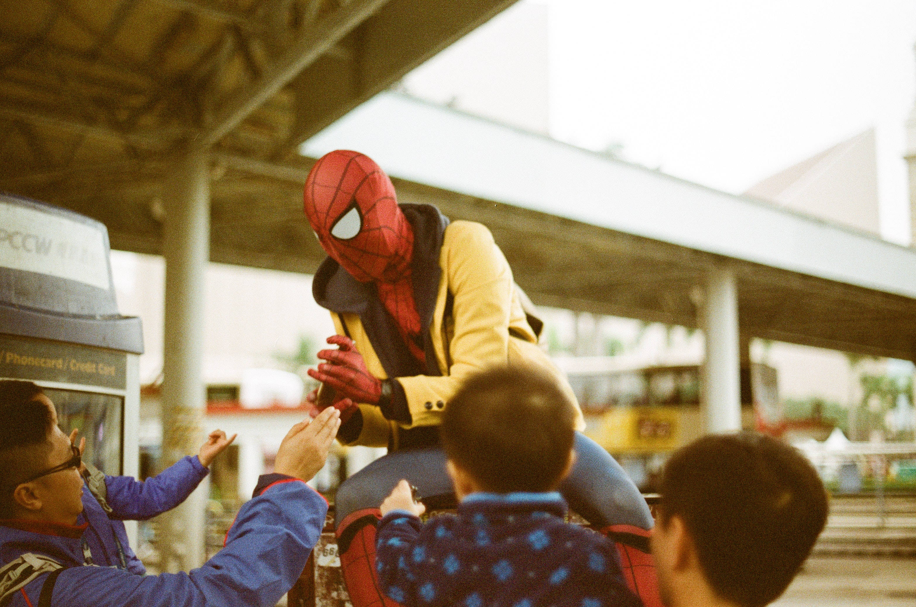 superhero supreme 桌布 壁紙 — 一名穿著蜘蛛人服裝的人在有蓋行人通道下的巴士站與兒童互動。
