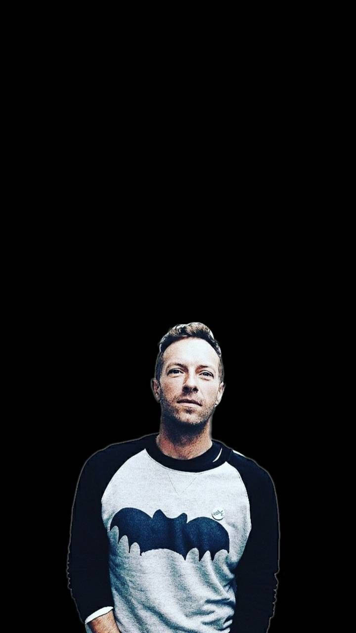 Chris Martin 桌布 壁紙 — 一名穿著蝙蝠俠標誌襯衫的男子站在純黑色背景前，燈光效果戲劇化。