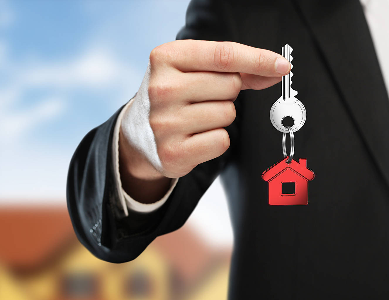 Luxury House 桌布 壁紙 — 一名穿著西裝的人手持一把帶有紅色房屋形標籤的銀色鑰匙，背景是模糊的郊區景色。
