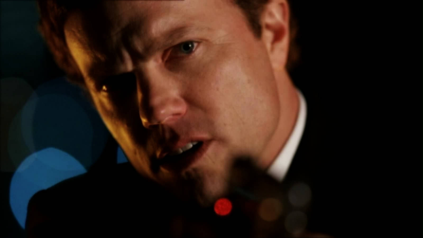 adam baldwin 桌布 壁紙 — 一名穿著西裝的男子在戲劇性燈光和模糊彩色散景背景下凝視著。