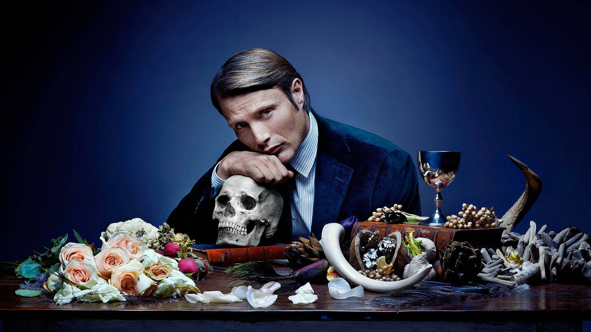 Hannibal 桌布 壁紙 — 一名穿著西裝的男子將頭靠在一個被玫瑰、角及骨頭包圍的骷髏頭上，背景為深藍色。