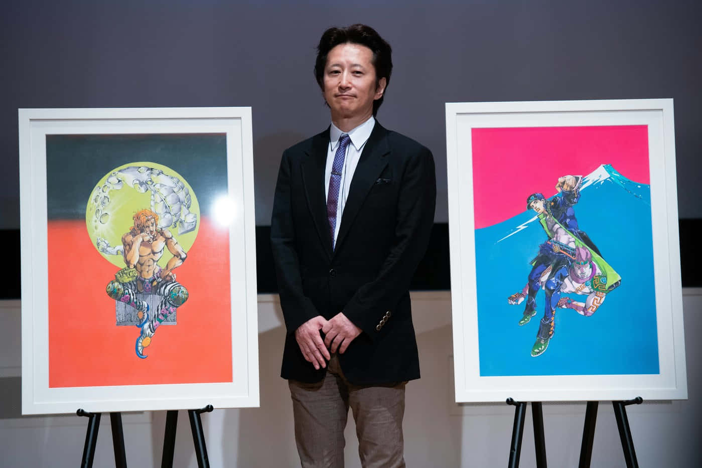 Hirohiko Araki 桌布 壁紙 — 一名穿著西裝的男子站在兩幅充滿活力的動漫風格裱框藝術品之間，這些藝術品以鮮豔色彩背景襯托出動態角色。