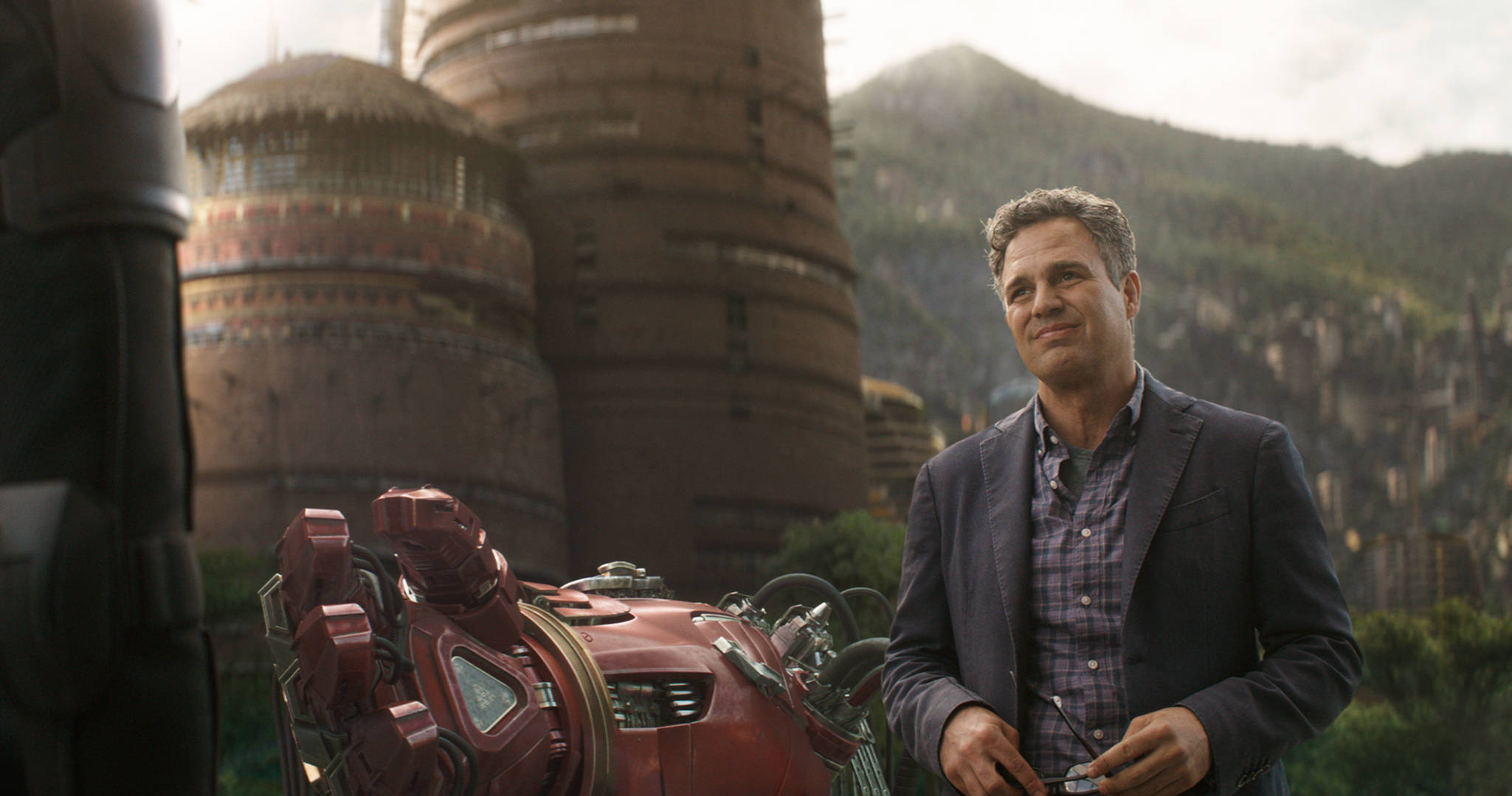 Mark Ruffalo 桌布 壁紙 — 一名穿著西裝的男子站在紅色裝甲服旁邊，背景是未來主義的塔樓和山脈城市景觀。