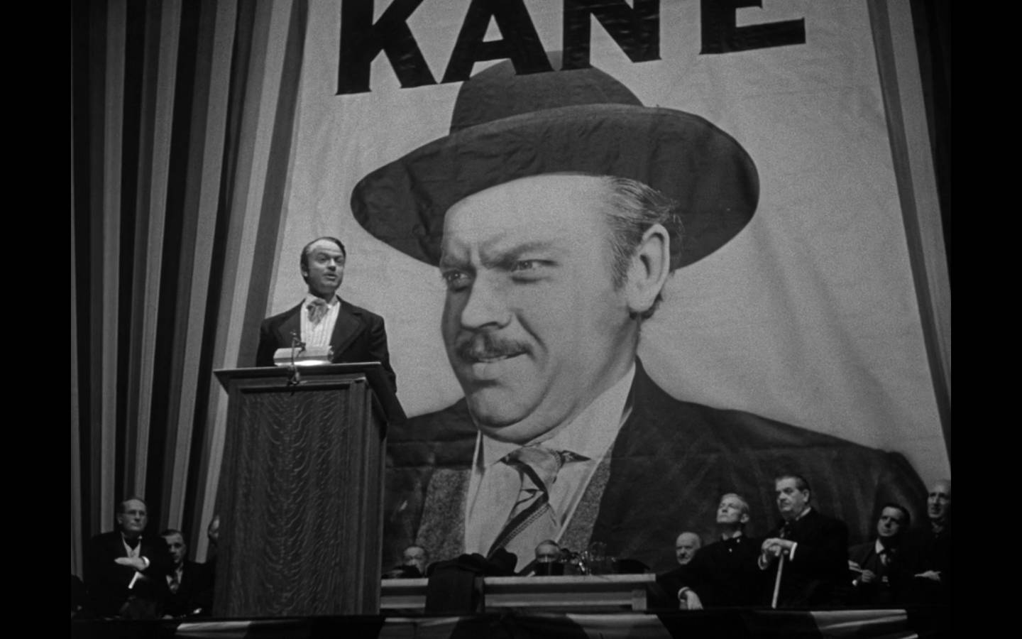 Citizen Kane 桌布 壁紙 — 一名穿著西裝的男子站在講台上，身後是一幅巨大的戴帽男子肖像，上面以粗體字顯示KANE這個名字。