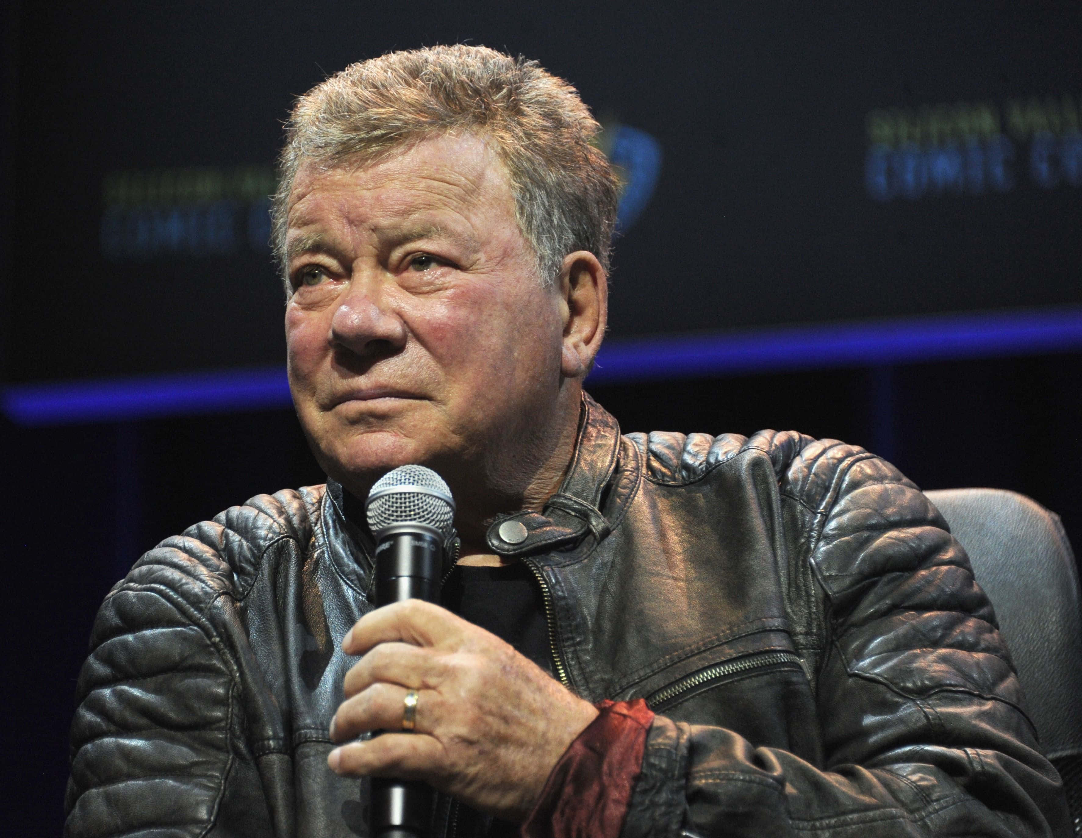 william shatner 桌布 壁紙 — 一名穿著質感皮革外套的男子在燈光昏暗、帶有藍色舞台燈光的活動中握著麥克風。