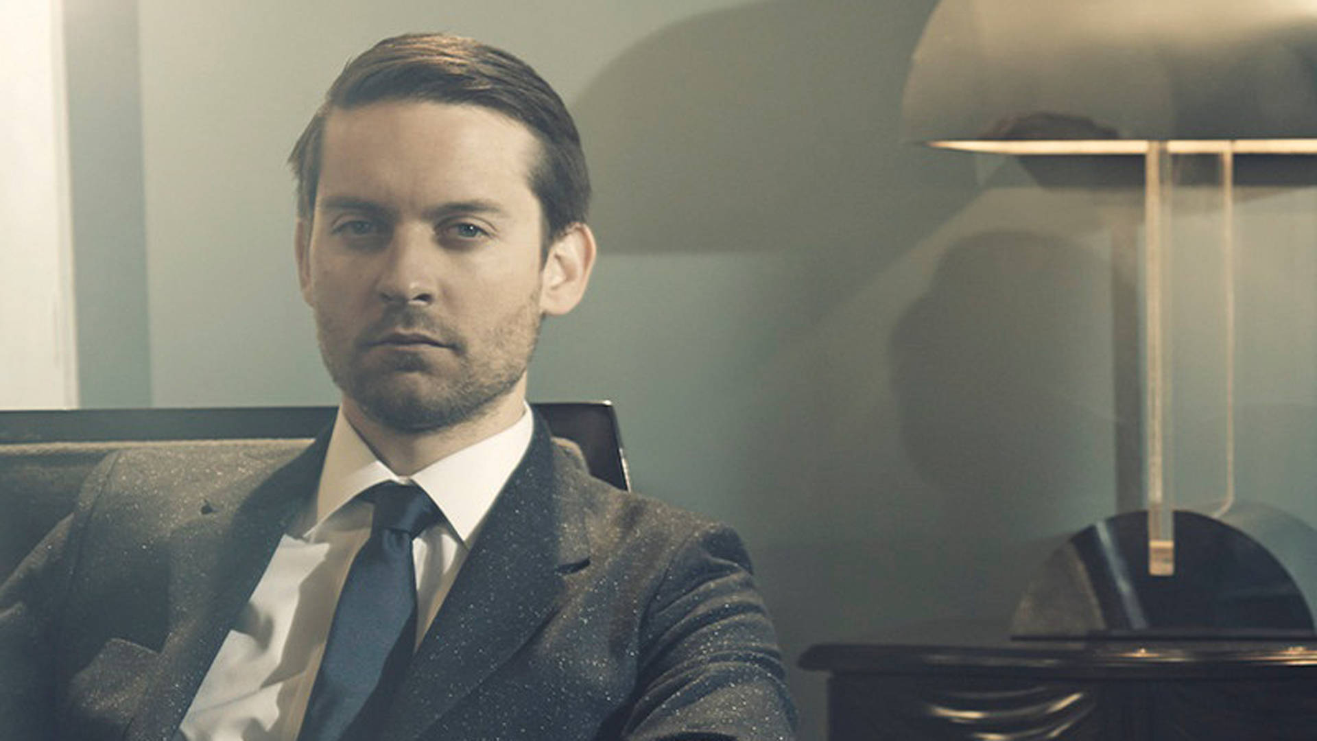 Tobey Maguire 桌布 壁紙 — 一名穿著質感西裝的男子坐在現代椅子上，旁邊是一盞時尚的燈，位於簡約風格的辦公室，環境光線柔和。