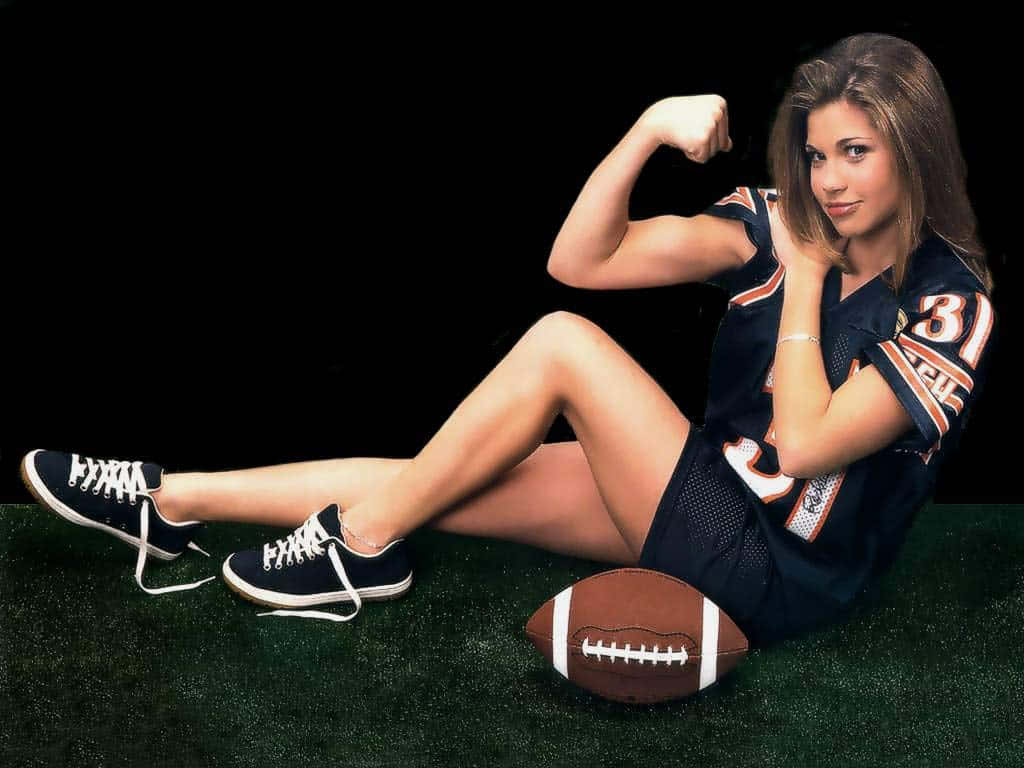 Danielle Fishel 桌布 壁紙 — 一名穿著足球球衣的女性坐在人造草地上，彎曲肱二頭肌，旁邊有一個足球。