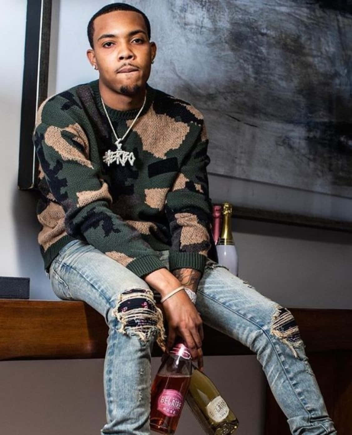 Herbo iPhone 桌布 壁紙 — 一名穿著迷彩毛衣和破洞牛仔褲的男子自信地坐著，手持一瓶玫瑰紅酒，背景是現代抽象牆。