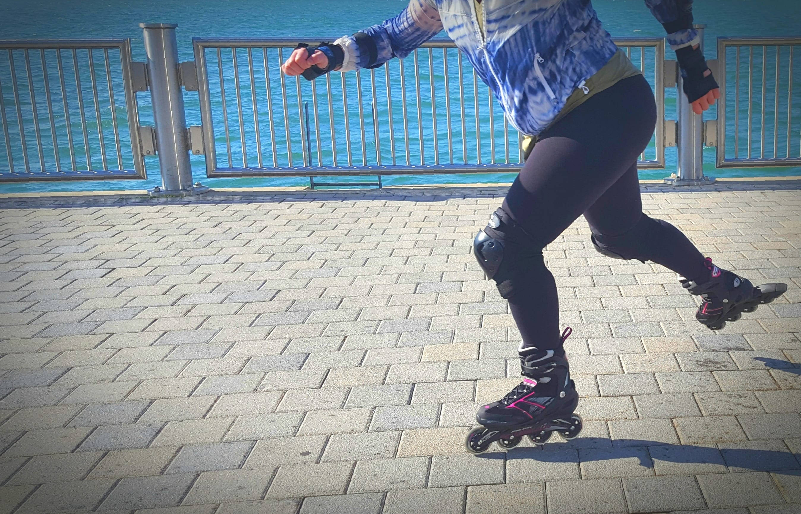 skating 桌布 壁紙 — 一名穿著防護裝備的人正在陽光普照的海濱長廊上溜直排輪，金屬欄杆後方是藍色海洋。