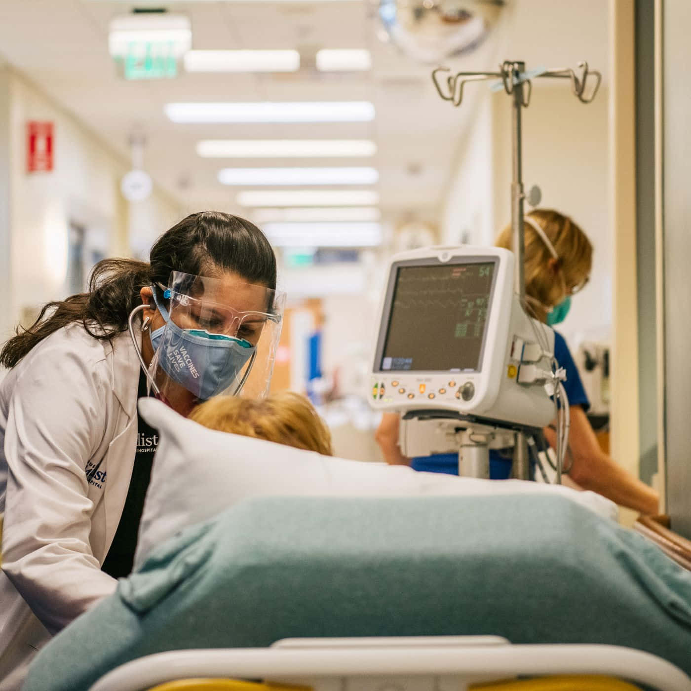 Nursing 桌布 壁紙 — 一名穿著防護裝備的護士俯身照顧醫院病床上的病人，同時在光線充足的走廊中，監視器顯示生命體徵。