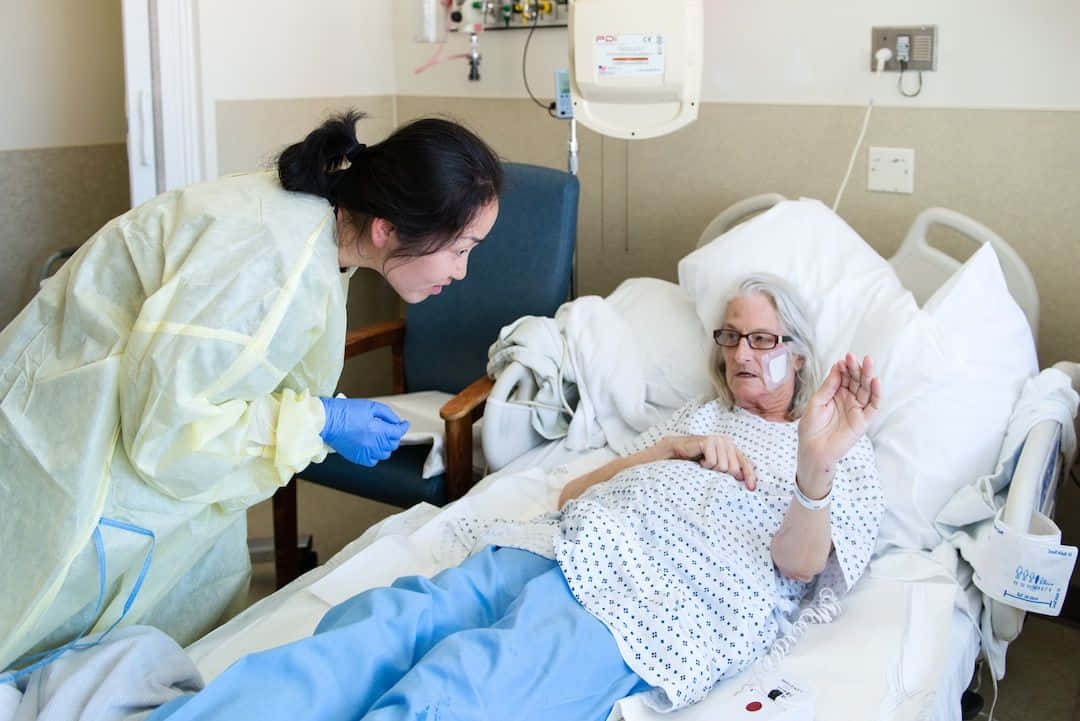 生病 桌布 壁紙 — 一名穿著防護裝備的護士正在照顧醫院病床上的一位老年病人，附近有醫療設備。