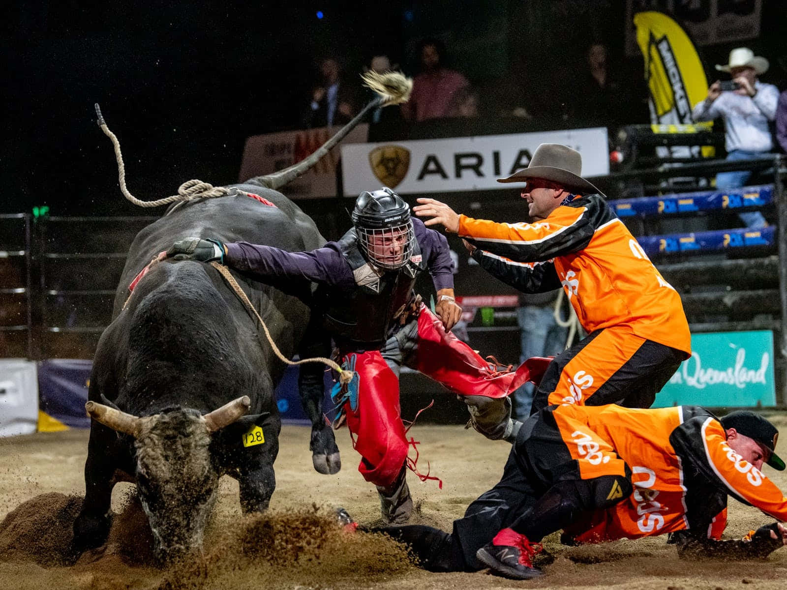 Bull Riding 桌布 壁紙 — 一名穿著防護裝備的騎牛士在夜間被一頭衝過來的黑牛拋下，兩名穿著橙色服裝的牛仔小丑在塵土飛揚的競技場上分散動物的注意力。