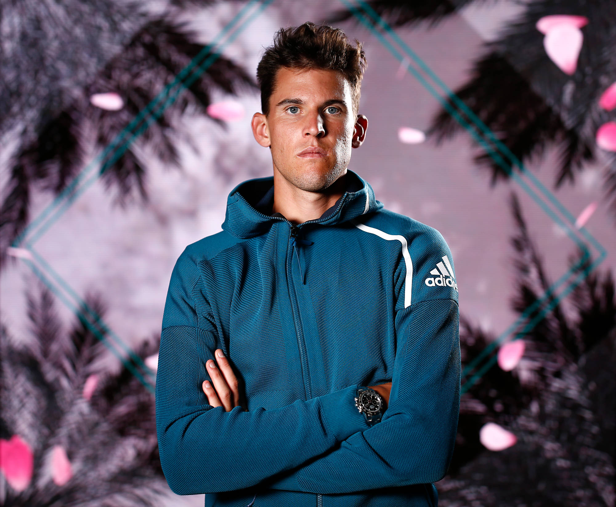 dominic thiem 桌布 壁紙 — 一名穿著青綠色Adidas連帽衫的男子，雙臂交叉，自信地站在一個風格化的棕櫚樹背景前，背景有粉紅色花瓣。