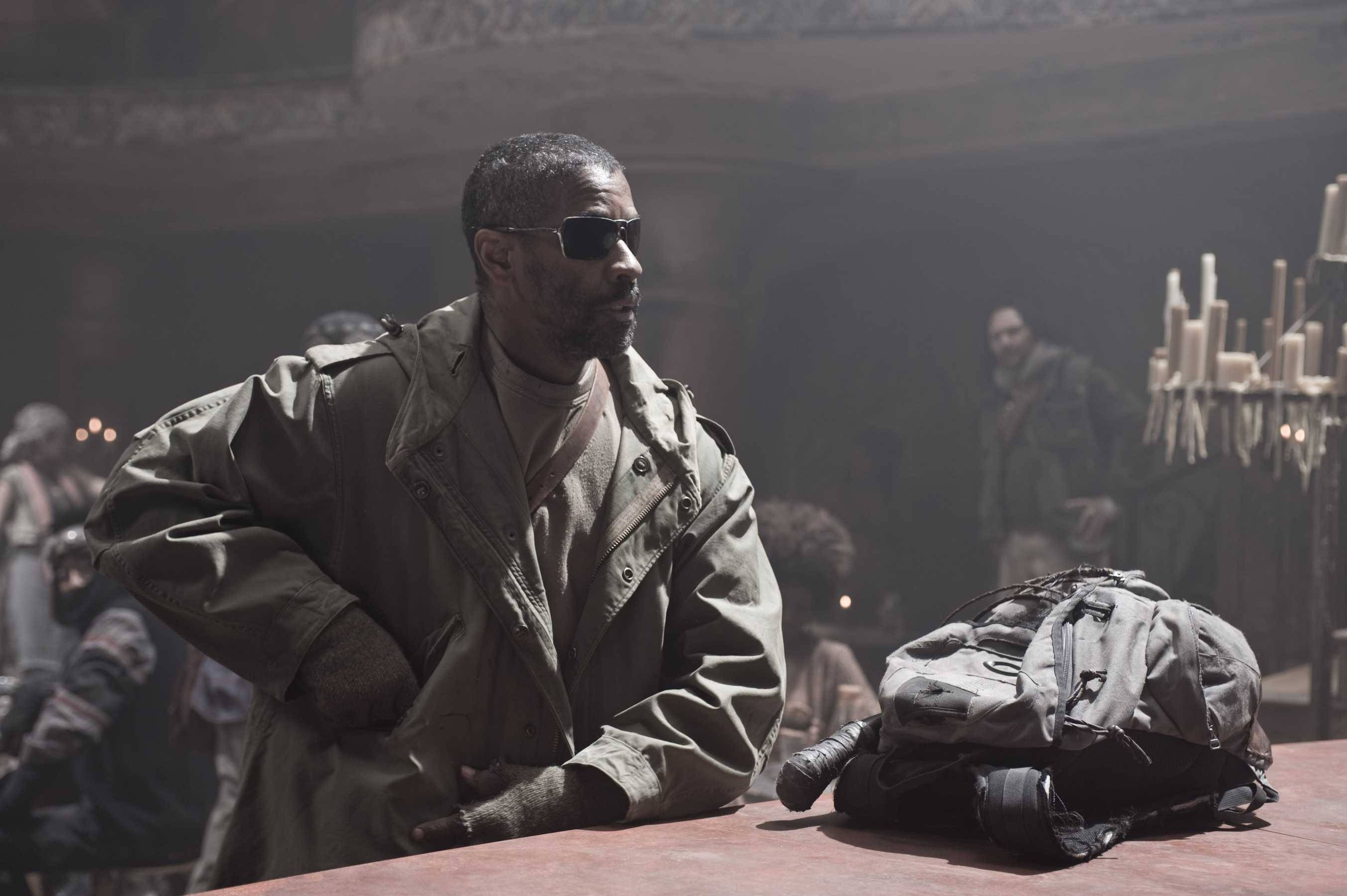 Denzel Washington 桌布 壁紙 — 一名穿著風衣和太陽眼鏡的男子靠在桌子上，旁邊放著相機包，身處燈光昏暗、煙霧瀰漫的房間。