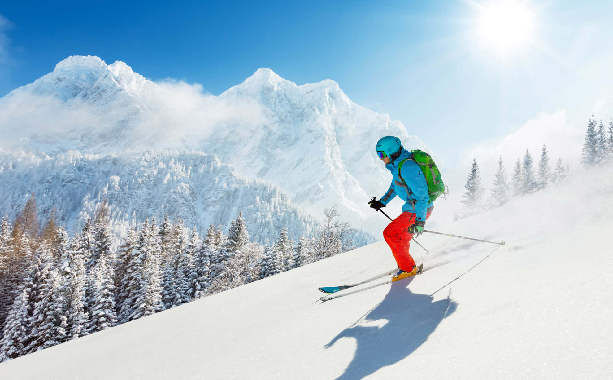 ski 桌布 壁紙 — 一名穿著鮮豔裝備的滑雪者，在晴朗藍天下，沿著白雪覆蓋的山坡滑行，背景是白雪覆蓋的山脈和常青樹。