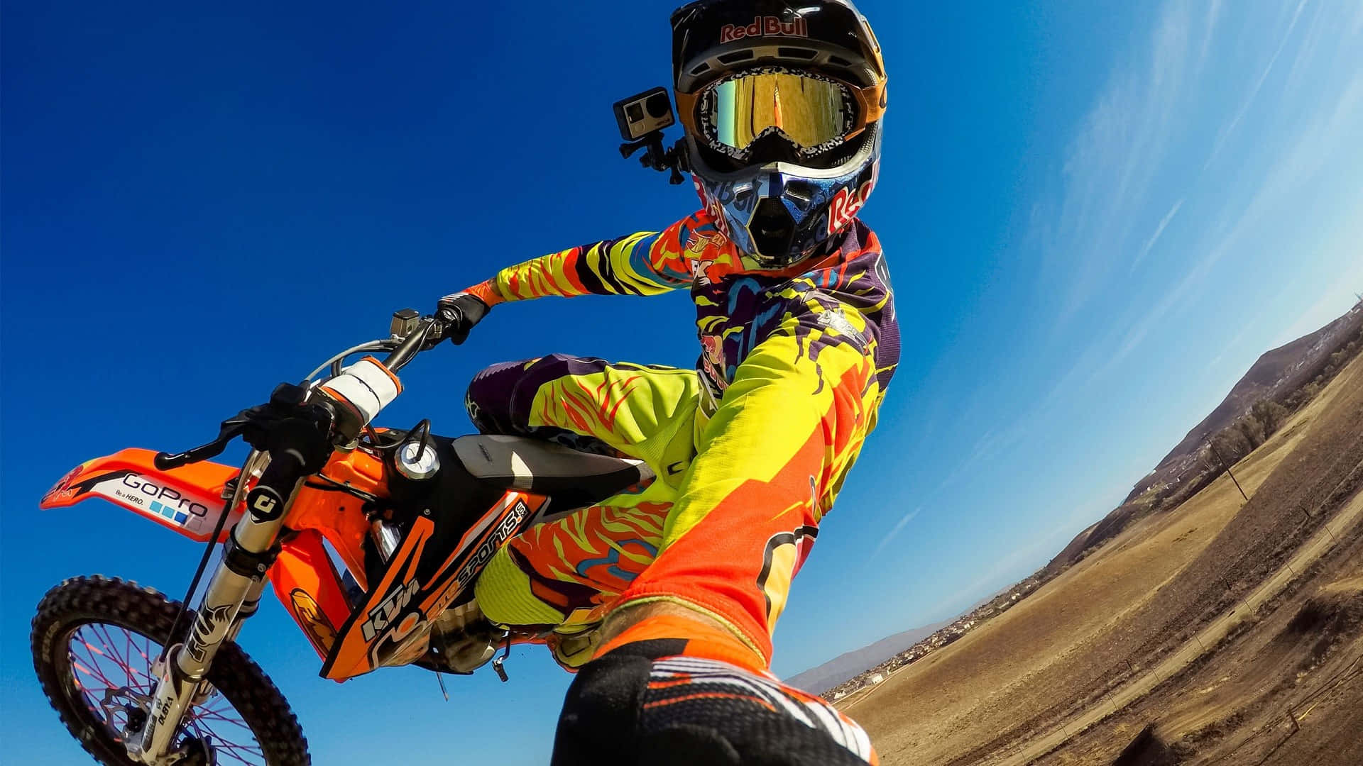 Motocross 桌布 壁紙 — 一名穿著鮮豔裝備的越野摩托車手在橙色越野車上傾斜入彎，背景是清澈的藍天。