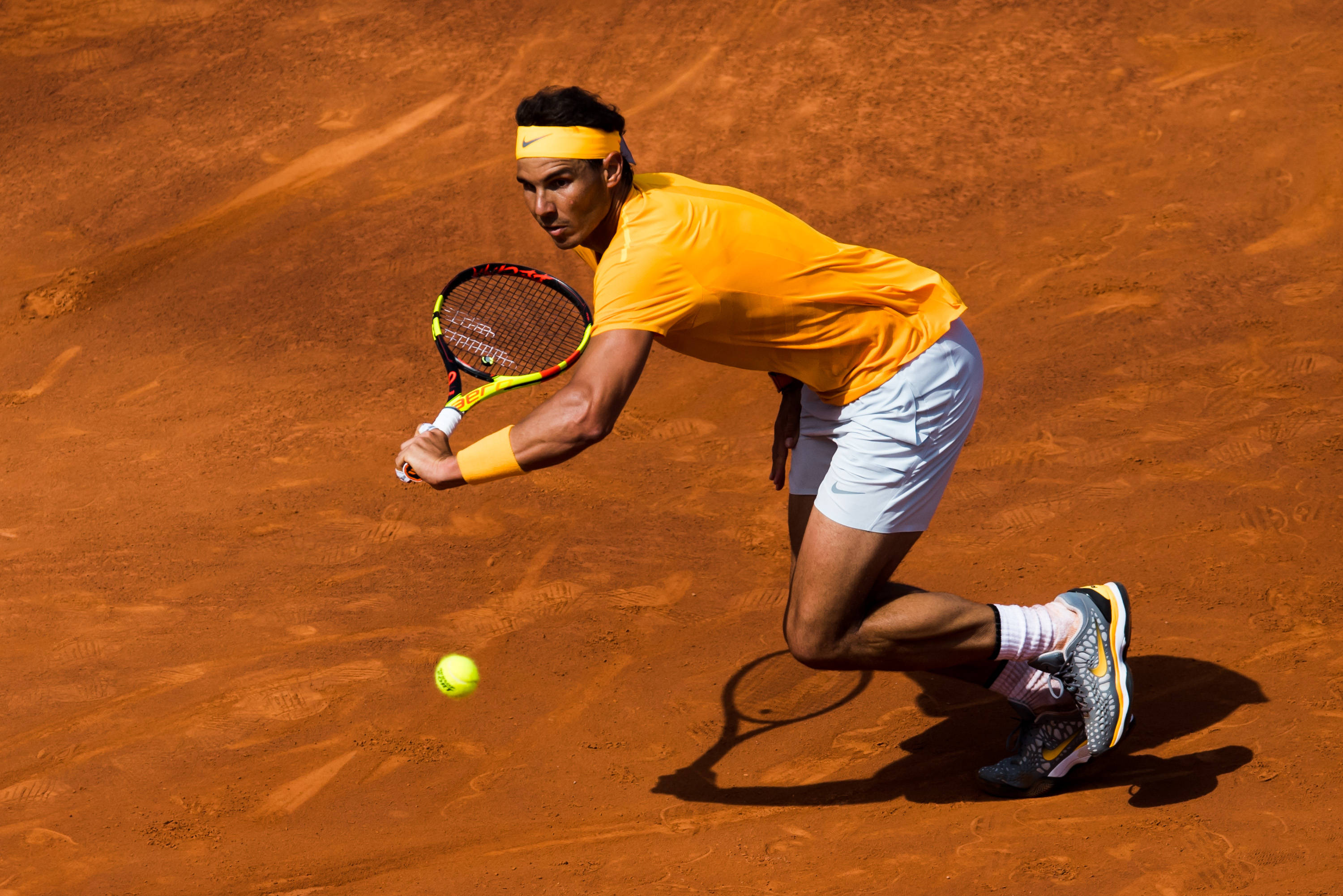 Tennis 桌布 壁紙 — 一名穿著黃色裝備的網球選手在紅色紅土球場上低身撲向一顆明亮的黃色球，在強烈的陽光下擊球。