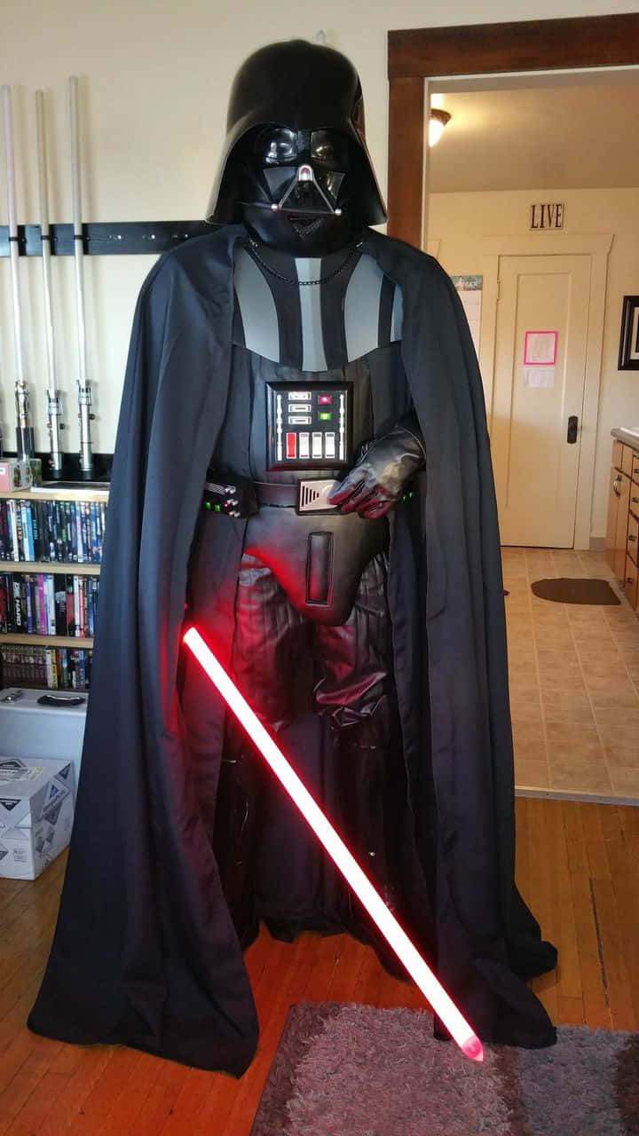 The Chosen One 桌布 壁紙 — 一名穿著黑武士服裝的人站在室內，在家中環境中手持發光的紅色光劍。