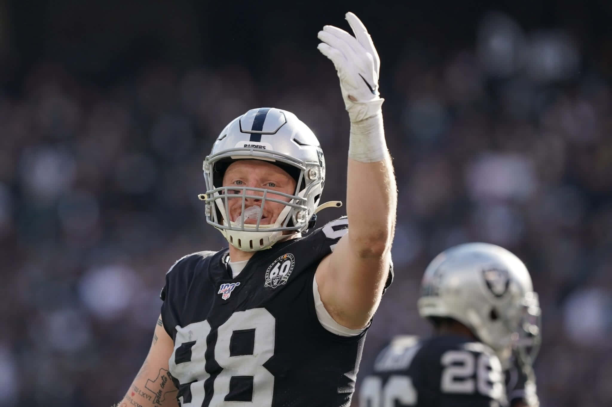 Las Vegas Raiders 桌布 壁紙 — 一名穿著黑色和銀色Raiders球衣的足球員，在陽光明媚的比賽日戴著頭盔和手套向球迷揮手。