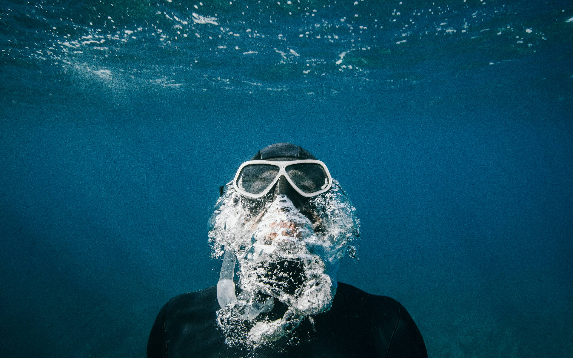 Swimming 桌布 壁紙 — 一名穿著黑色潛水衣和白色面罩的潛水員在水下呼氣，在深藍色海洋背景下產生一陣泡沫狀的氣泡。