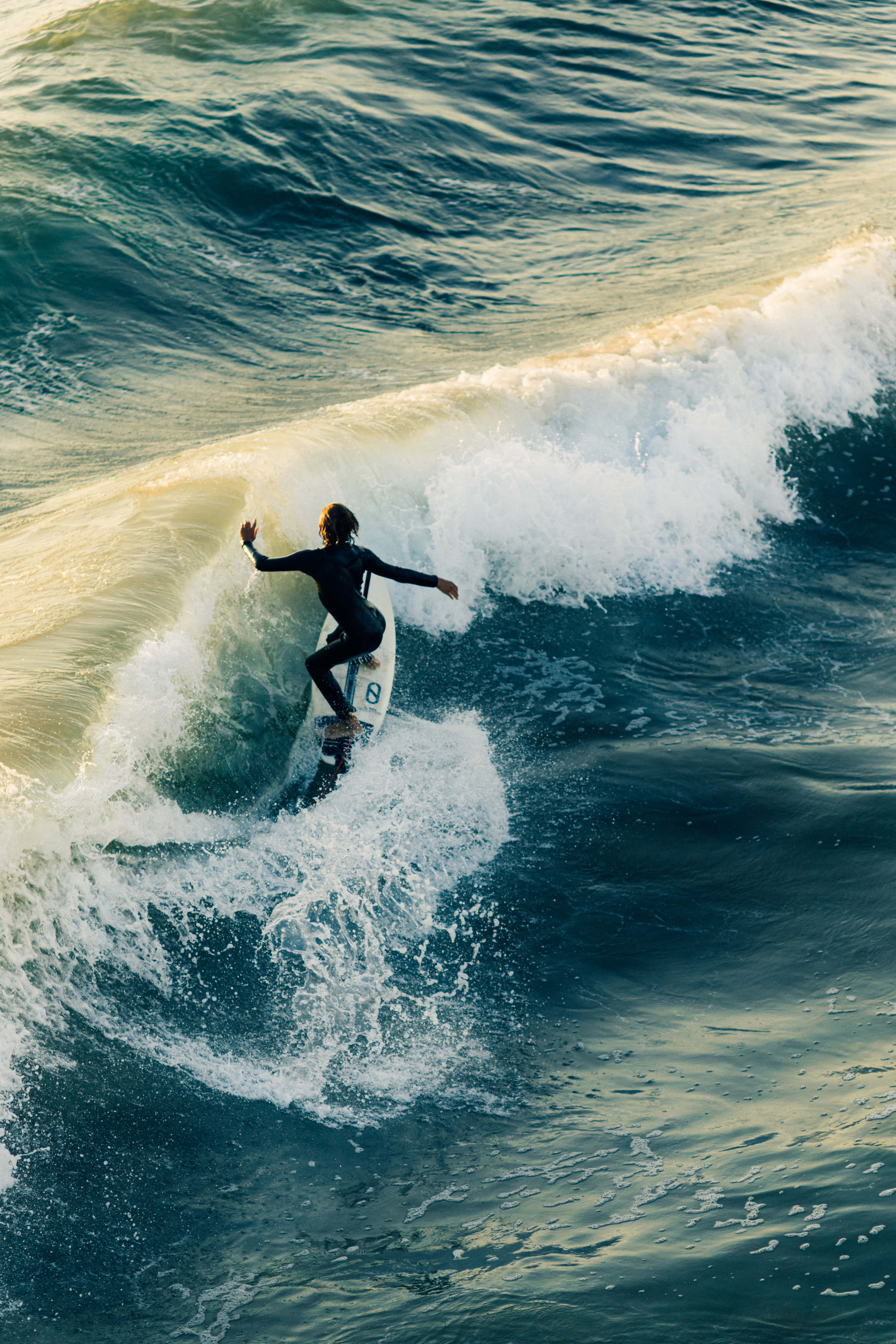 Surfing iPhone 桌布 壁紙 — 一名穿著黑色潛水衣的衝浪者，雙臂伸展，在深藍色的海水中駕馭強勁的海浪。