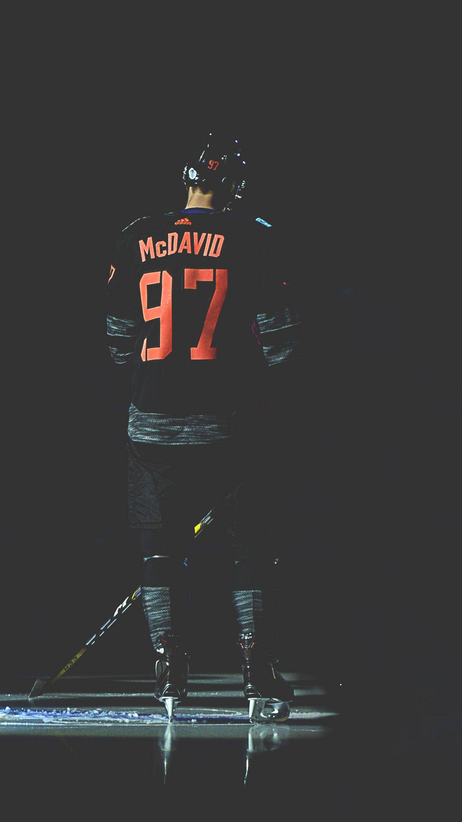 hockey 桌布 壁紙 — 一名穿著黑色球衣、帶有橙色McDavid 97的冰球運動員獨自站在冰場上，處於戲劇性的聚光燈下。