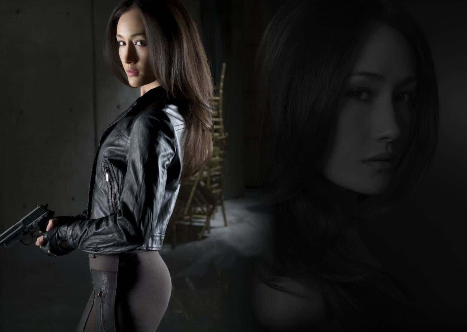 maggie q 桌布 壁紙 — 一名穿著黑色皮夾克的女子在光線昏暗的工業空間中手持槍支，並帶有鬼臉疊加效果。