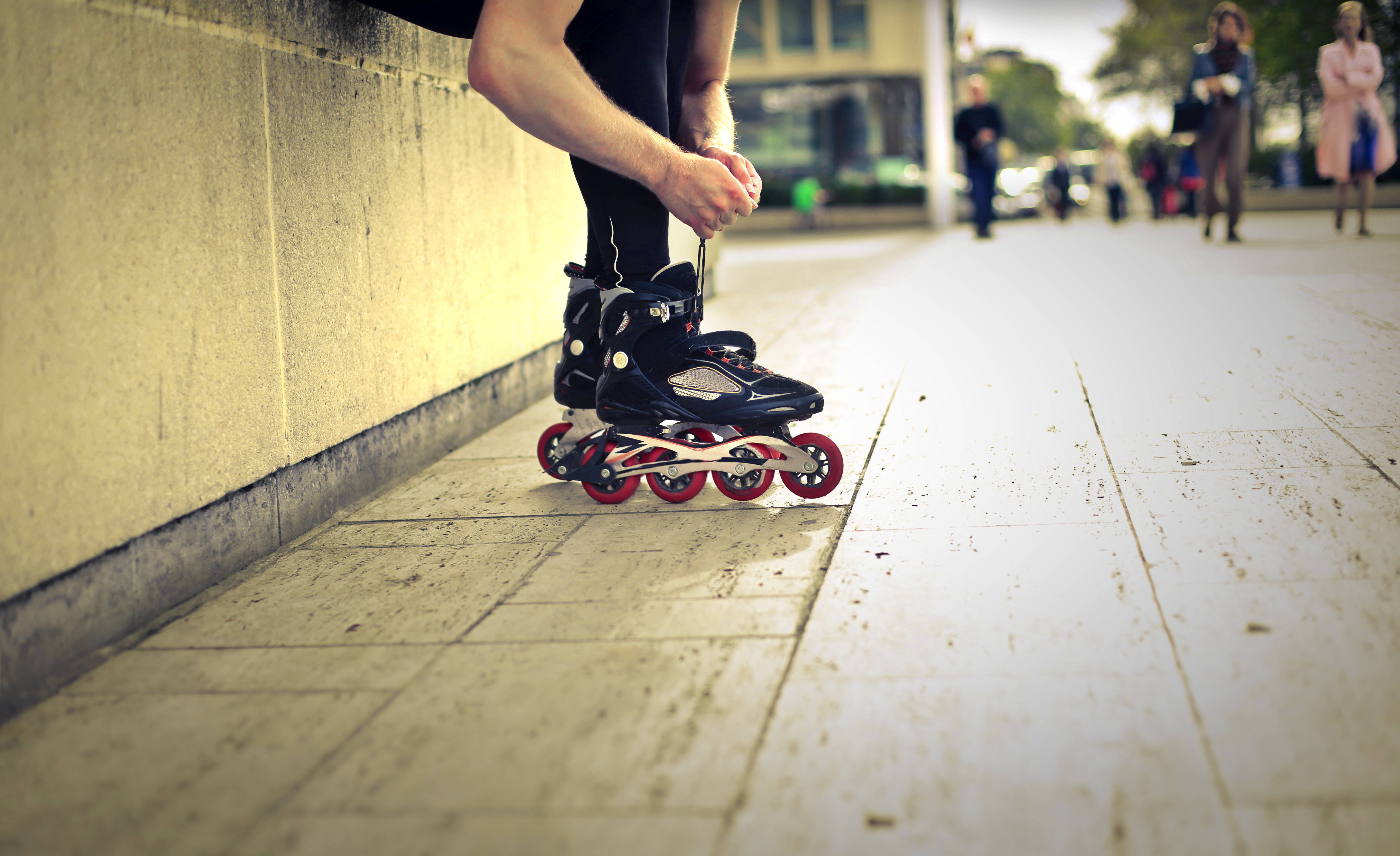skating 桌布 壁紙 — 一名穿著黑色緊身褲的人正在陽光普照的城市人行道上，靠近混凝土牆調整帶有紅色輪子的直排輪。