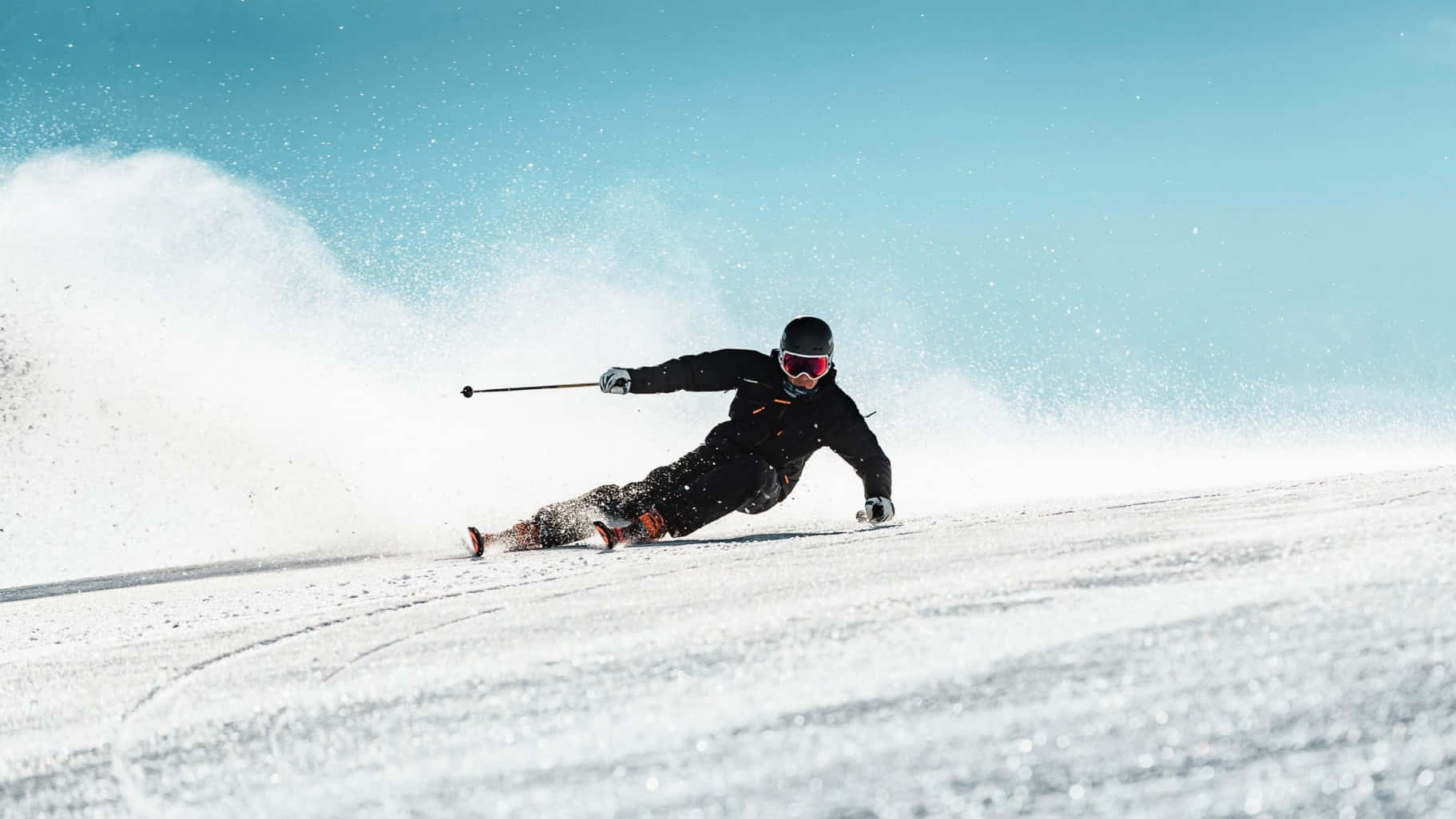 ski 桌布 壁紙 — 一名穿著黑色裝備的滑雪者在雪坡上急轉彎，將粉雪揚起至明亮的藍天。