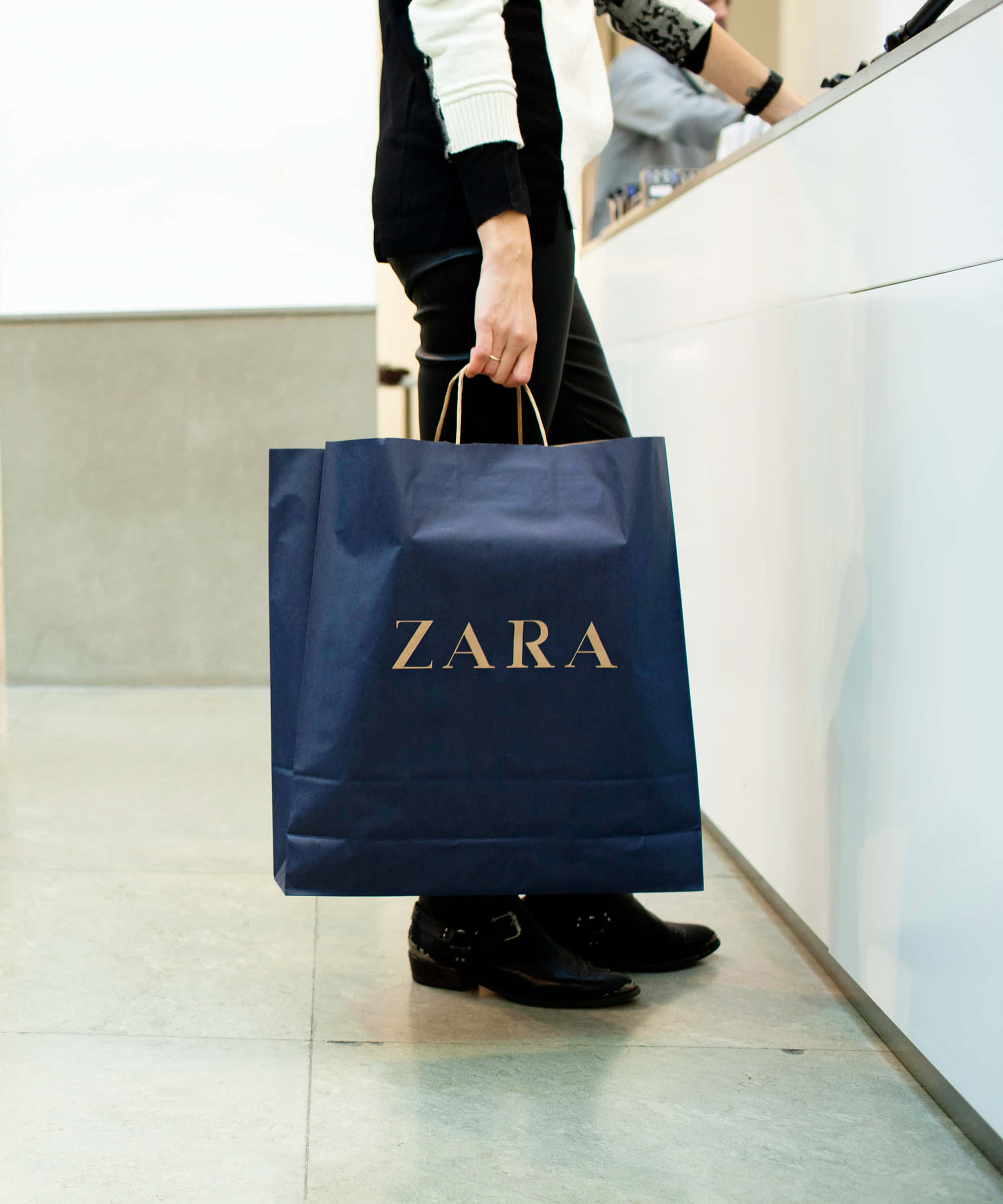 Zara 桌布 壁紙 — 一名穿著黑色褲子和靴子的人，在現代零售櫃檯前拿著一個深藍色Zara購物袋。