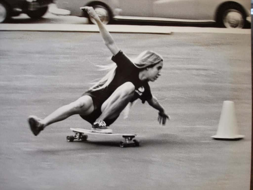 Retro Skateboard 桌布 壁紙 — 一名穿著黑色襯衫和短褲的滑板手在鋪砌的停車場上低身滑過一個圓錐，背景中有模糊的汽車。