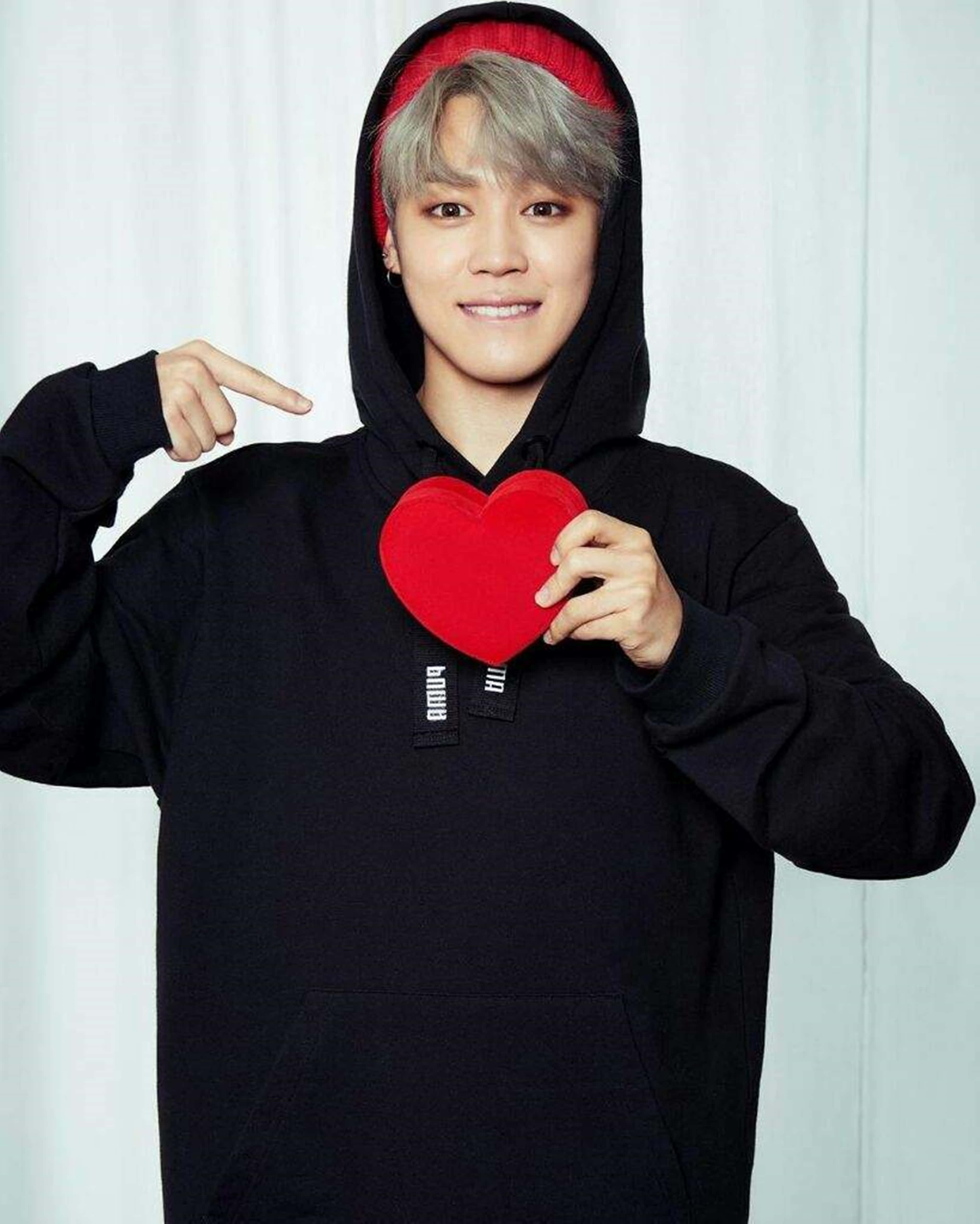 Bts Jimin 桌布 壁紙 — 一名穿著黑色連帽衫（帽子內襯紅色）的年輕男子，在白色窗簾背景下，手持一個紅色心形物體並指向它。