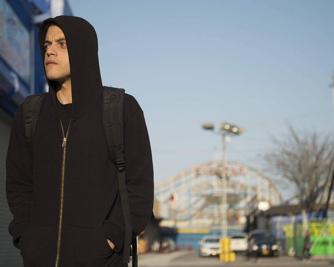Rami Malek 桌布 壁紙 — 一名穿著黑色連帽衫的年輕男子在晴朗的藍天下沉思地站在戶外，背景是模糊的嘉年華。