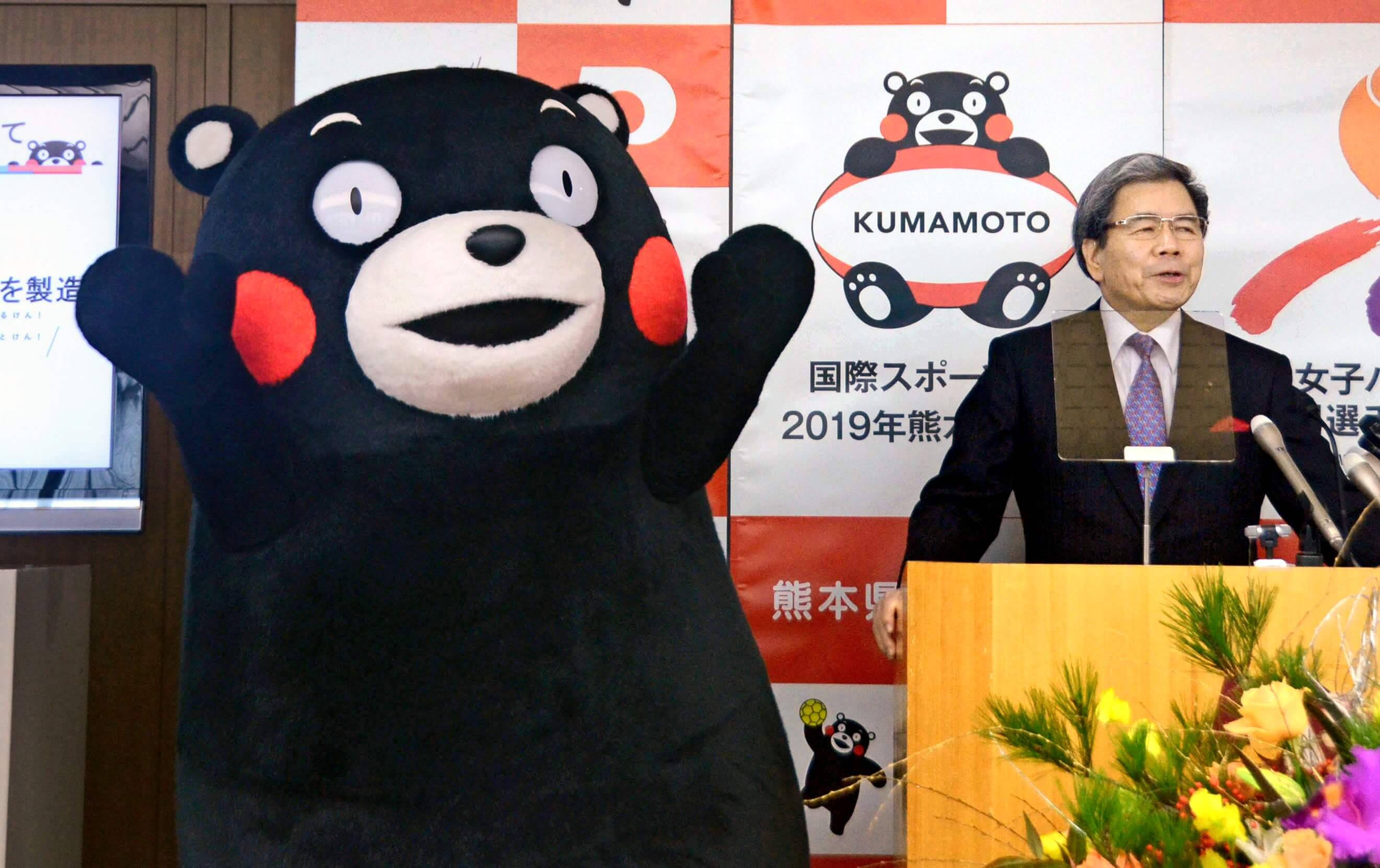 熊本熊 桌布 壁紙 — 一名穿西裝的男子在活動中於講台旁發言，旁邊有一個大型 Kumamon 吉祥物在揮手。
