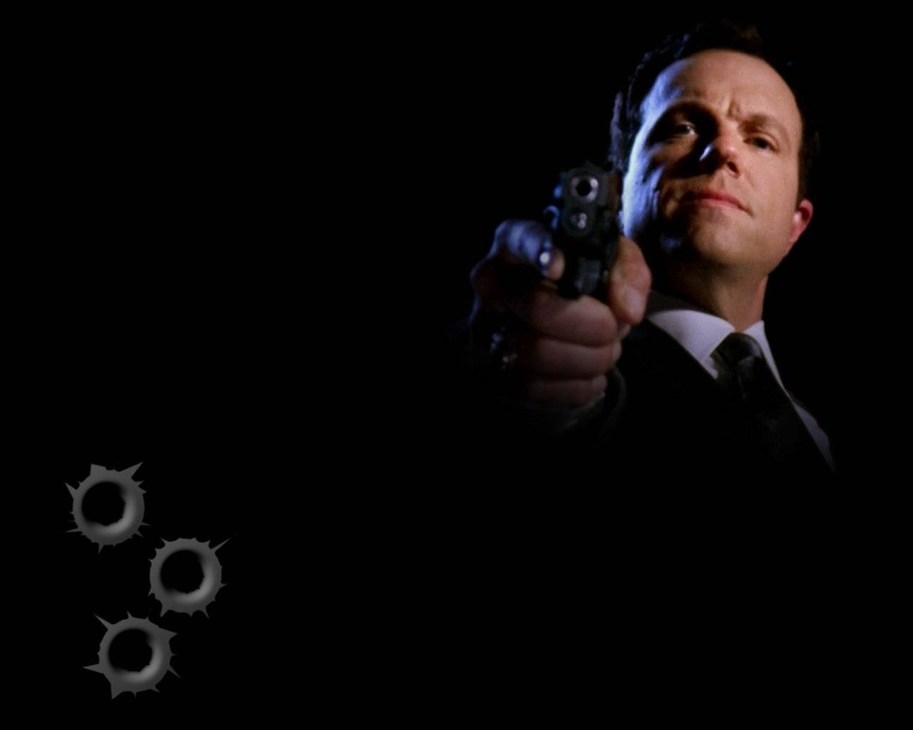 adam baldwin 桌布 壁紙 — 一名穿西裝的男子在黑色背景前直接用手槍瞄準觀眾，左下角有三個彈孔。