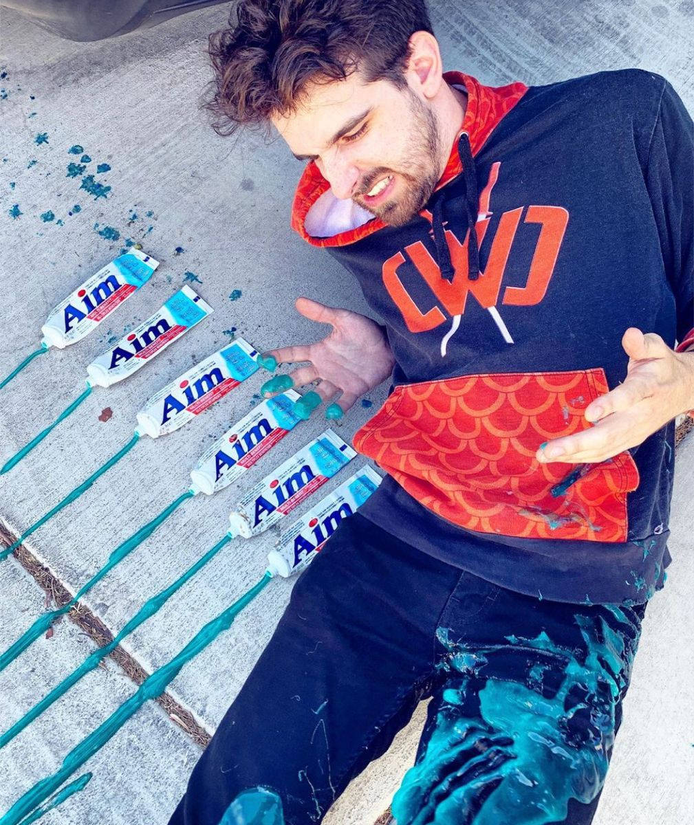 Man 桌布 壁紙 — 一名穿連帽衫的男子躺在人行道上，身上覆蓋著從多支Aim牙膏管擠出的青綠色牙膏。