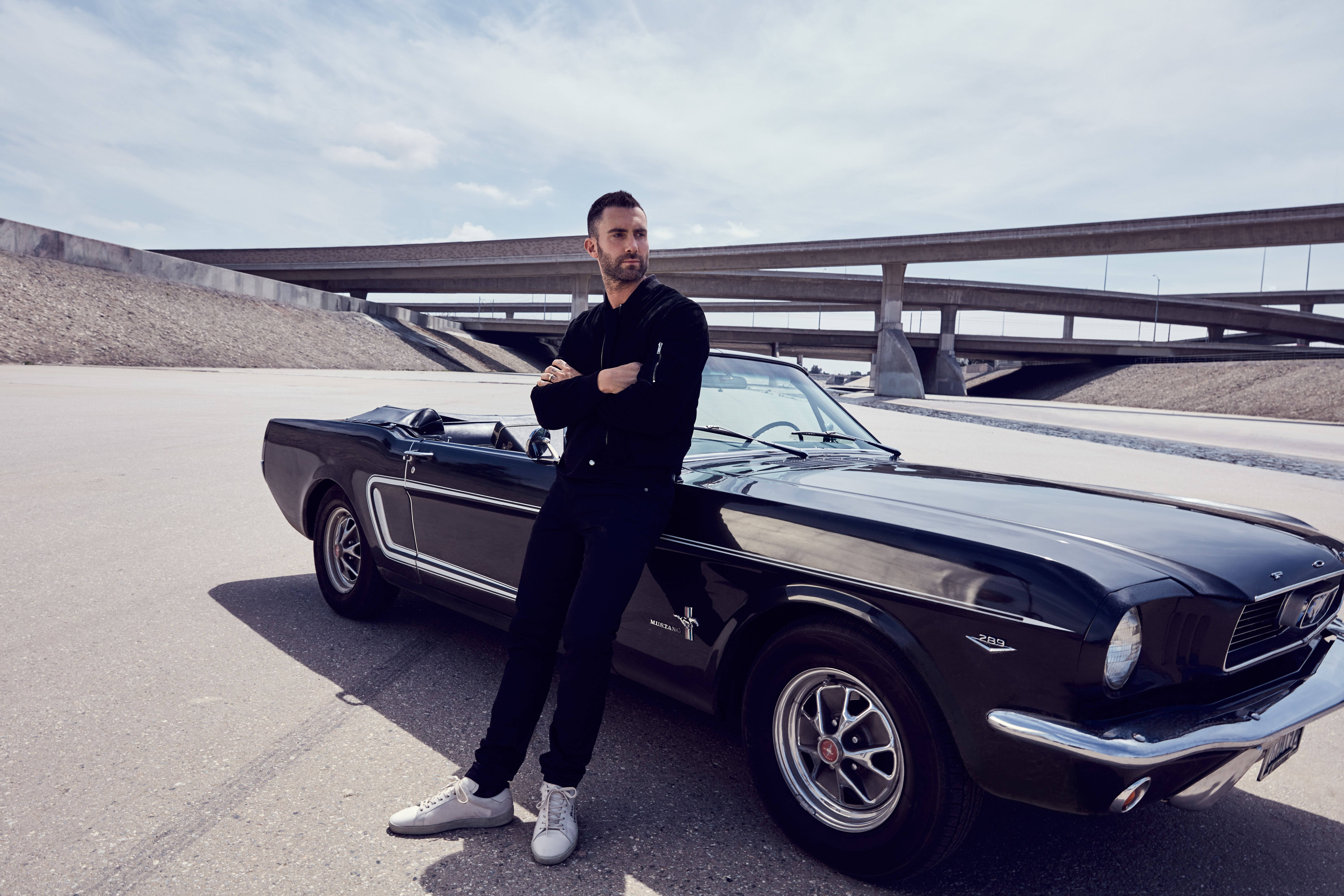 Adam Levine 桌布 壁紙 — 一名穿黑衣的男子自信地靠在一輛閃亮的黑色經典Mustang敞篷車旁，背景是廣闊的天空和混凝土高架橋。
