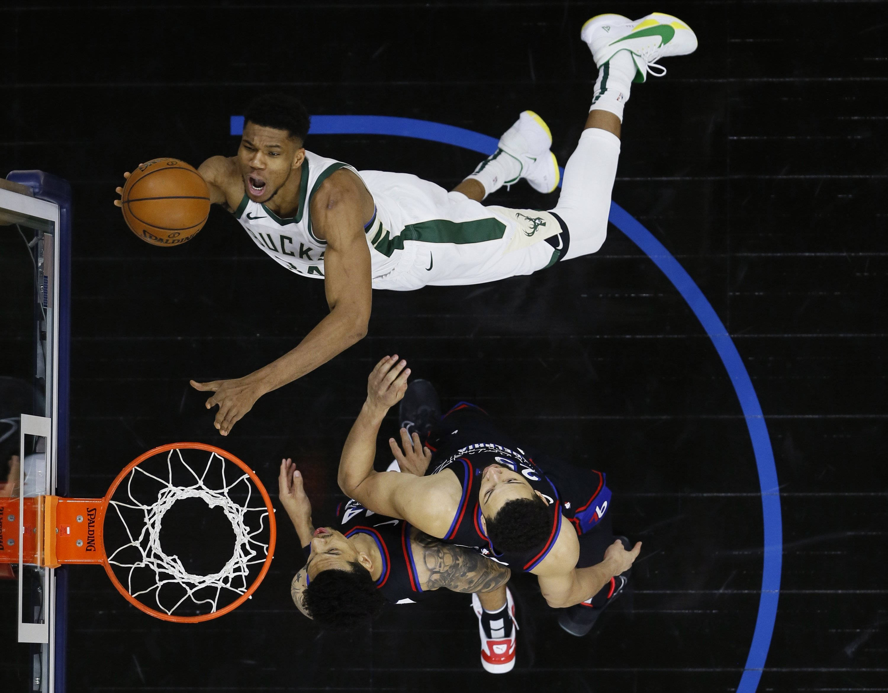 Giannis 桌布 壁紙 — 一名籃球運動員在半空中灌籃，而防守球員在他下方跌倒在黑暗的球場上。