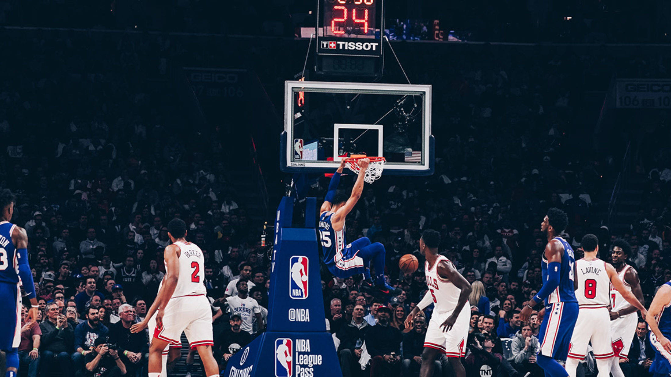 76ers 桌布 壁紙 — 一名籃球運動員在半空中灌籃，隊友和對手在場館燈光下觀看。