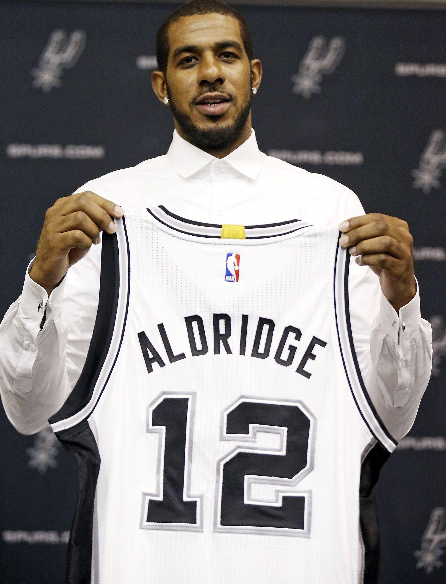 Lamarcus Aldridge 桌布 壁紙 — 一名籃球運動員舉起一件白色Spurs球衣，上面有Aldridge和12號，背景為深藍色。
