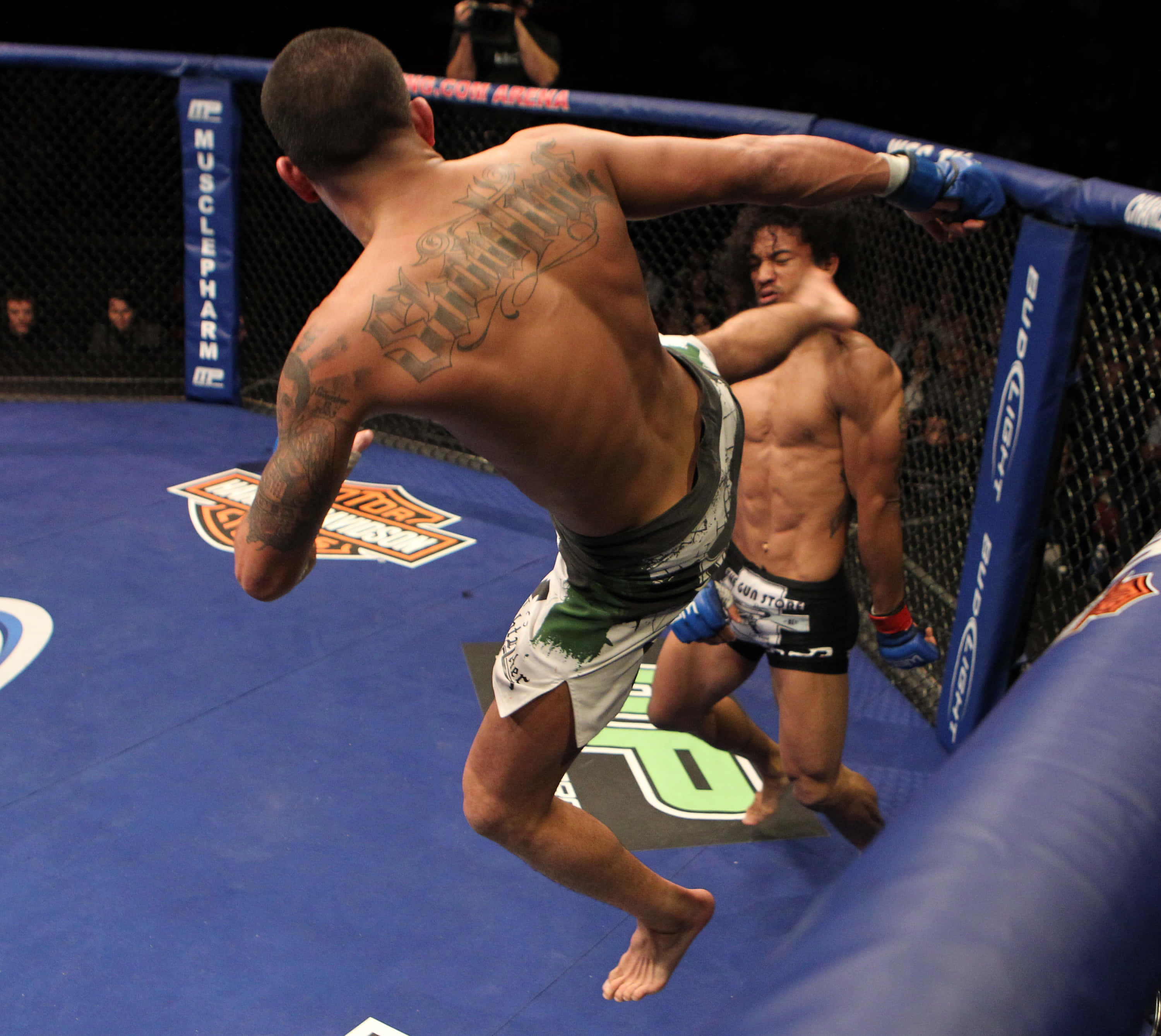 MMA 桌布 壁紙 — 一名紋身拳手在籠中躍起，於綜合格鬥比賽中向對手踢出強力一腳。