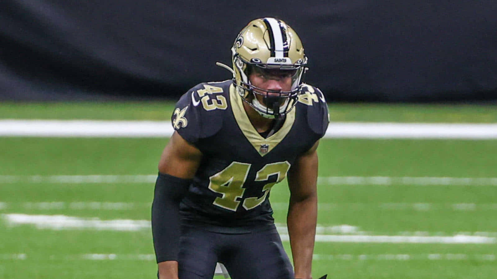 Marcus Williams 桌布 壁紙 — 一名紐奧良聖徒隊的美式足球員穿著金色和黑色制服，在體育場燈光下的綠色球場上激烈地蹲伏。