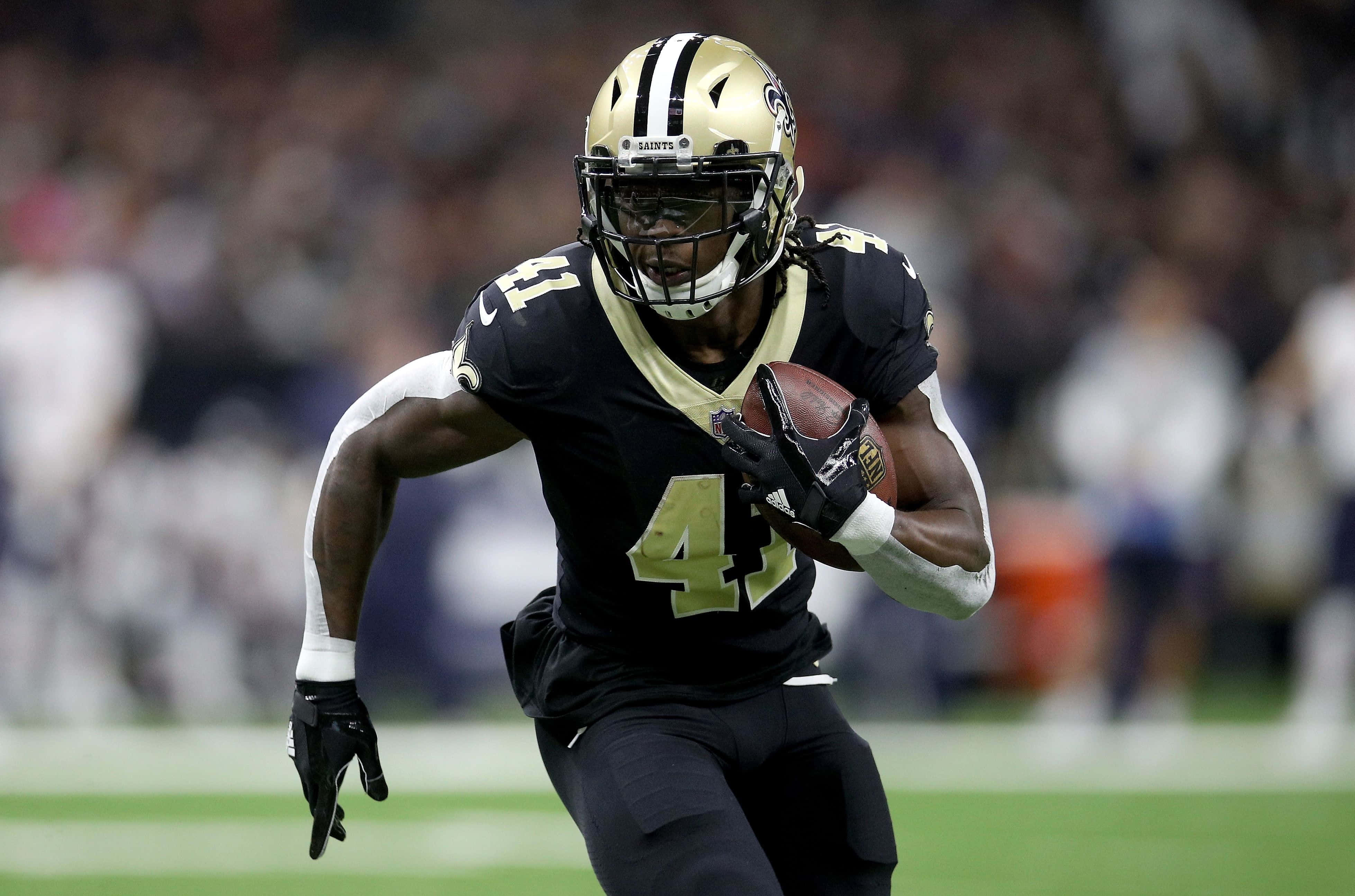 Alvin Kamara 桌布 壁紙 — 一名紐奧良聖徒隊美式足球員身穿黑金色球衣，在球場燈光下比賽期間持球衝刺。