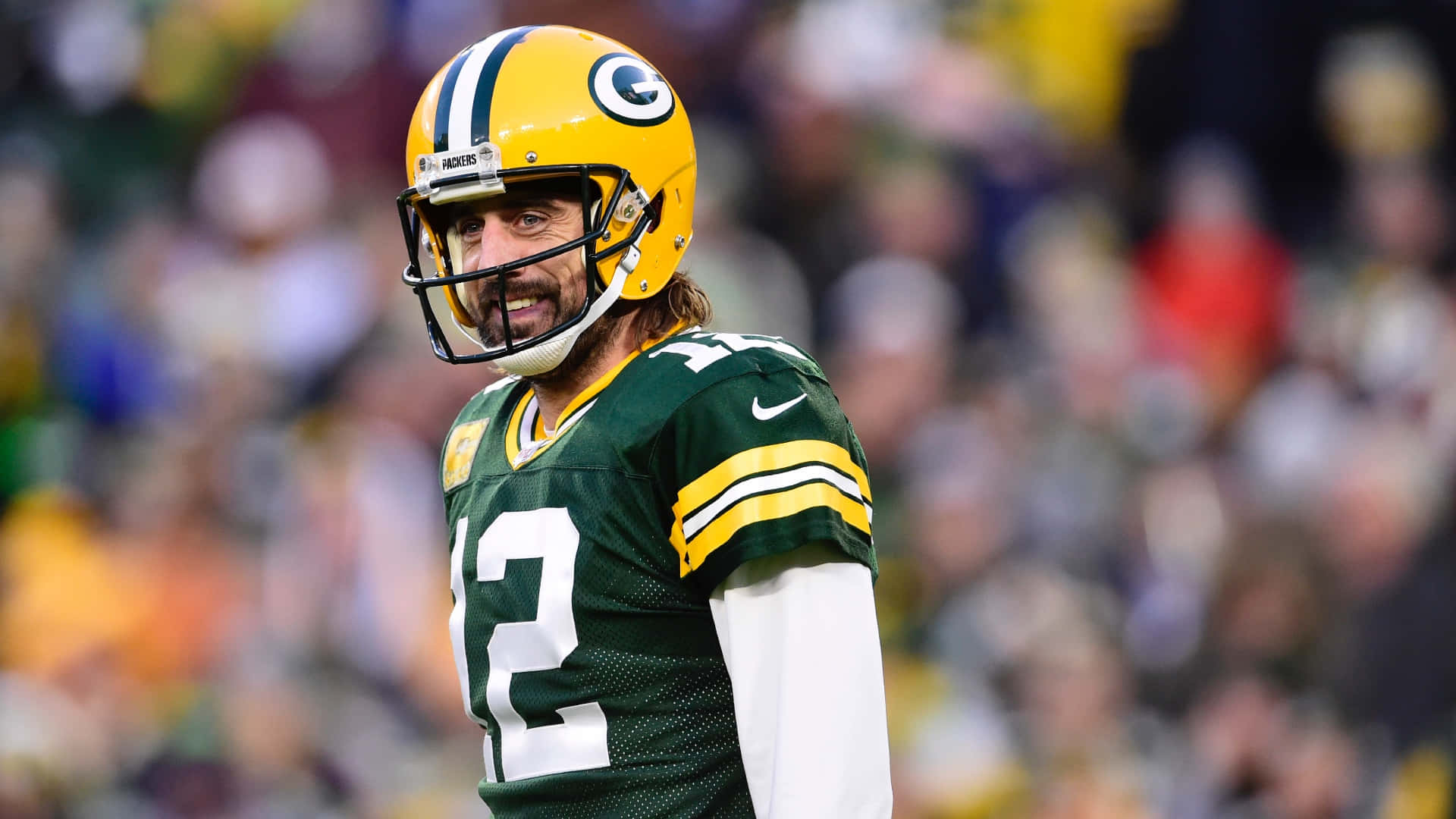 Aaron Rodgers 桌布 壁紙 — 一名綠灣包裝工美式足球員身穿綠色和金色制服，專注地站在球場上，身後是模糊的體育場人群。