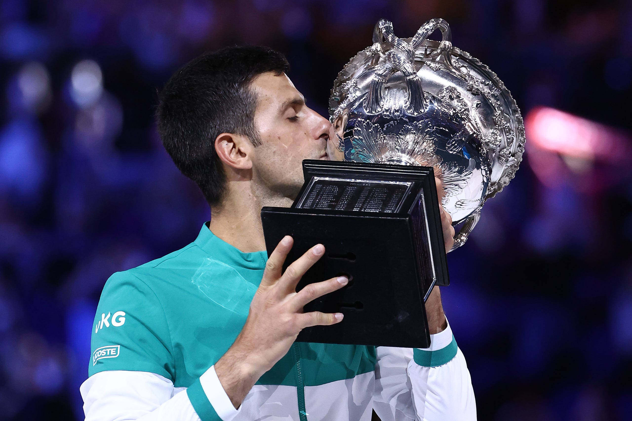 Australian Open 桌布 壁紙 — 一名網球運動員在明亮的體育場燈光下親吻一個大型銀色獎盃，身後是模糊的人群。