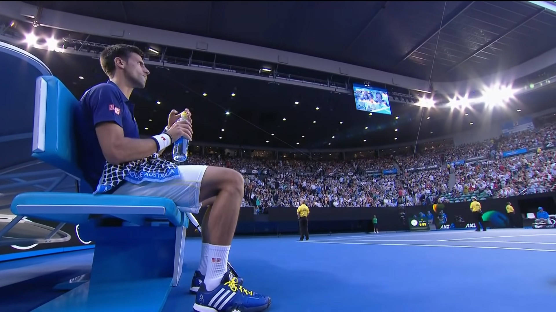 Novak 桌布 壁紙 — 一名網球運動員坐在充滿活力的藍色球場的藍色長椅上，手持水壺，座無虛席的體育館在明亮的燈光下閃耀。