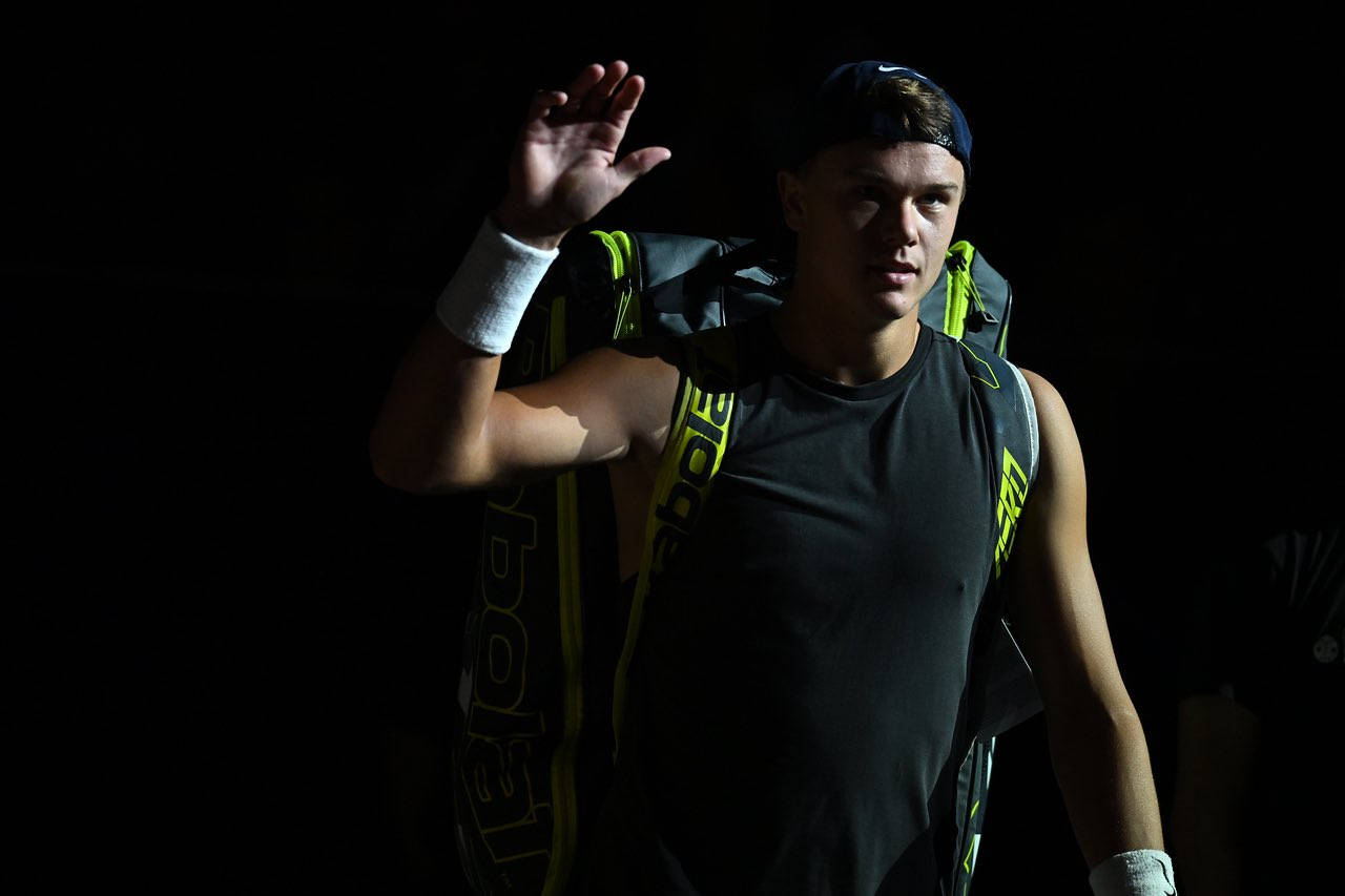 Tennis 桌布 壁紙 — 一名網球運動員帶著專注的表情揮手，同時背著一個亮黃色和黑色的球拍袋，背景為深色。