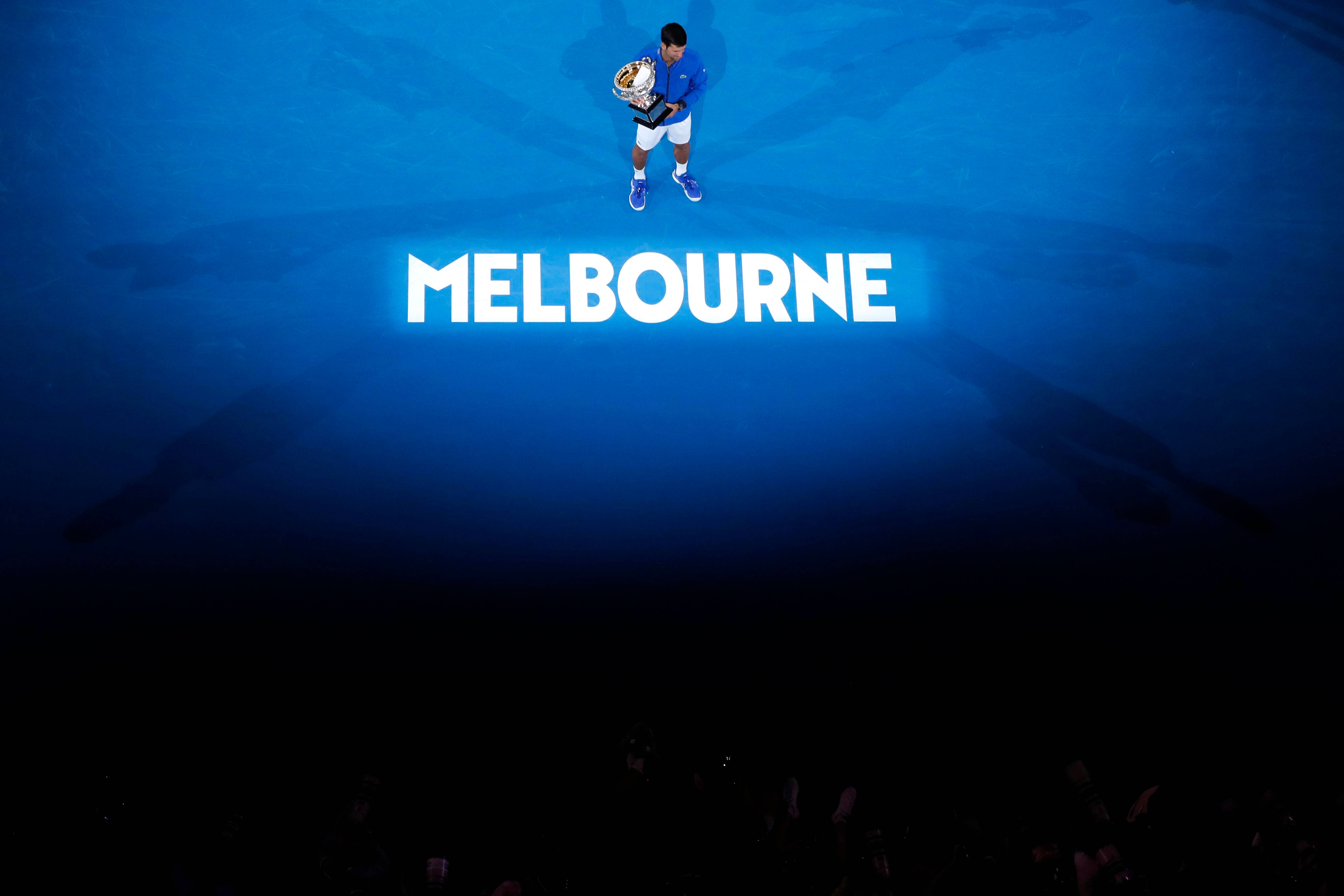 Australian Open 桌布 壁紙 — 一名網球選手獨自站在藍色球場上，在明亮的燈光下高舉獎盃，而「MELBOURNE」字樣在他腳下閃耀。