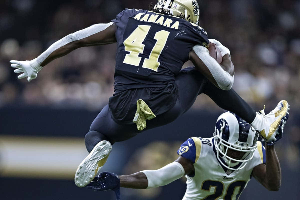 Alvin Kamara 桌布 壁紙 — 一名美式足球員在半空中跳起緊抱球，而防守員伸手試圖擒抱他，這是在夜間比賽中發生的。