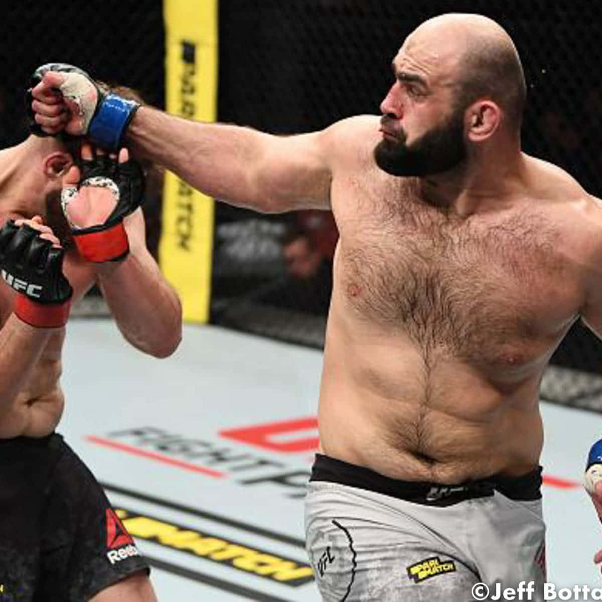 UFC 桌布 壁紙 — 一名肌肉發達、光頭留鬍的拳手在UFC八角籠內進行綜合格鬥比賽時打出強力一拳。